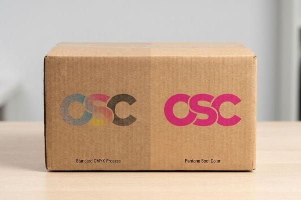 Brown corrugated box displaying CSC logos: left in faded Standard CMYK Process, right in vibrant Pantone Spot Color for brand accuracy.