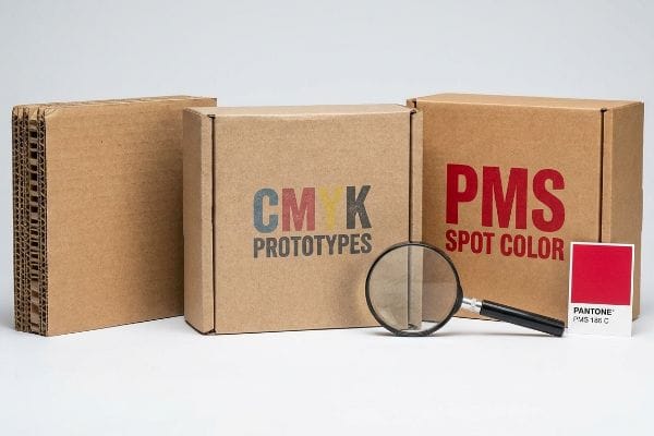 CMYK PMS Printing Comparison Corrugated cardboard boxes display Cmyk Prototypes and Pms Spot Color printing, with a magnifying glass and Pantone Pms 186 C swatch.
