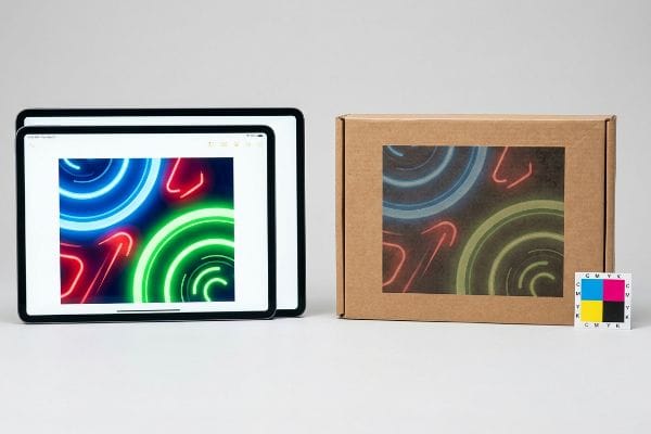 Color Model Comparison Digital tablets display vibrant neon art, contrasted with the same design printed on a cardboard box, next to a CMYK color chart.