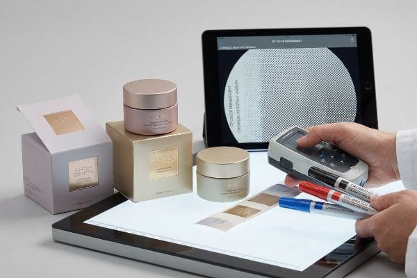 Colorimeter measures cosmetic packaging, tablet shows G7 Color Management and Chemical-Resistant Varnish, with Calibrated dyne pens.