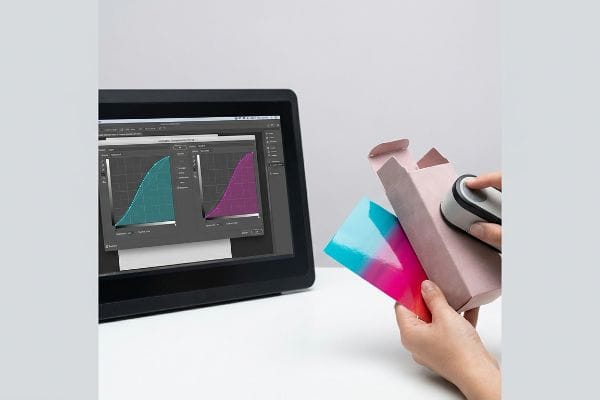 A colorimeter scans soft-touch packaging, displaying Lamination Compensation Curves on a monitor for color accuracy.
