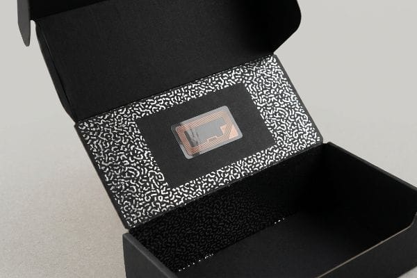 Etiqueta RFID para embalaje inteligente Etiqueta RFID transparente con bobinas de antena de cobre integradas en la tapa negra de una caja de embalaje inteligente abierta, con un patrón moteado plateado.