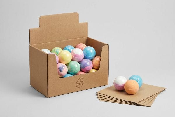 Bath Bomb Display Packaging Brown corrugated cardboard display box filled with pastel bath bombs, featuring a 'Sustainable Packaging' logo, next to flat brown sheets.