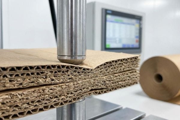 Corrugated Board Strength Test Multiple layers of brown corrugated paperboard with visible flutes undergoing a compression test, assessing structural strength for packaging.