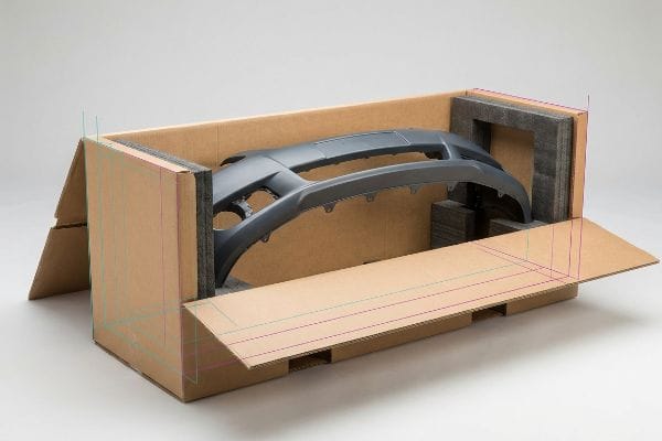Car Bumper Shipping Box An open custom corrugated shipping box, featuring faint CMYK registration lines, securely cradles a gray car bumper protected by dark foam inserts for robust automotive part shipping.
