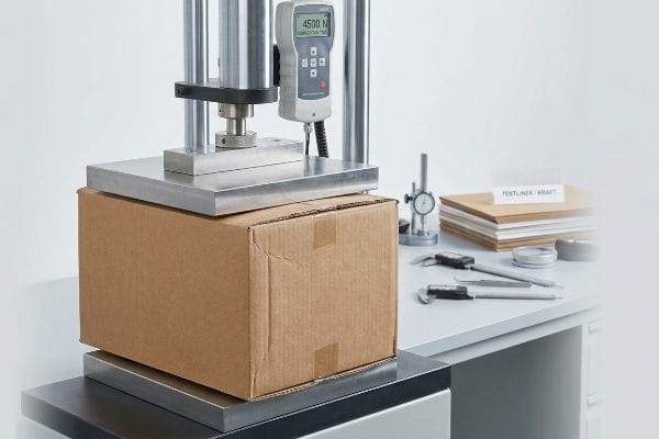 Brown corrugated box undergoing compression testing, a digital force gauge shows 4500 N, with Testliner / Kraft samples.