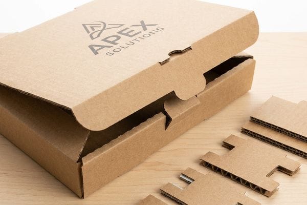 Apex Solutions Die-Cut Box An open brown corrugated die-cut box with Apex Solutions logo, featuring visible fluting and separate cardboard inserts on a wood surface.
