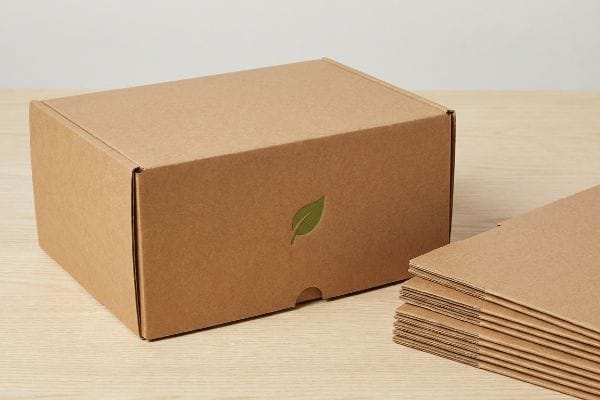 Uma caixa de papelão ondulado marrom com um logotipo de folha verde está ao lado de uma pilha de folhas planas de embalagem ecológica.