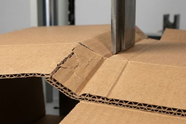 Cardboard Crease Stress Corrugated cardboard showing micro-fractures and buckling inner flutes as a steel rule die presses to create a fold.