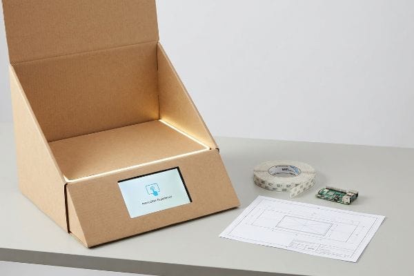 Display Tech Integration A corrugated display box with an integrated digital screen displaying 'Interaktive Experience' and LED lighting, shown with a circuit board, tape, and blueprints for technology integration.