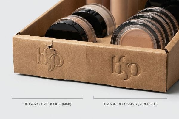 Embossing Debossing Comparison Corrugated merchandising tray comparing damaged Outward Embossing (Risk) with intact Inward Debossing (Strength) of the bsp logo, holding makeup compacts.