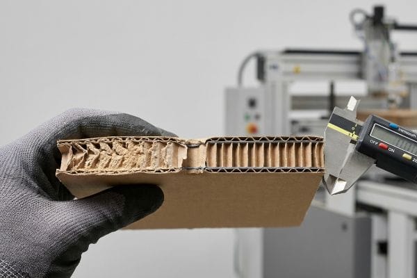 Gloved hand measures corrugated cardboard thickness with a digital caliper, showing crushed and intact flutes.