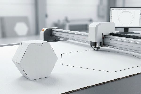 Digital Hexagon Box Cutting White corrugated cardboard hexagonal box and a Kongsberg digital cutting table precisely scoring a new hexagon design, with the design on a monitor.