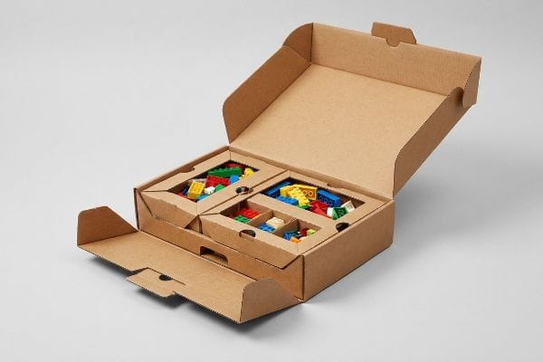 Brown corrugated cardboard box with open lid, revealing mono-material paperboard dividers holding colorful Lego-style building blocks.