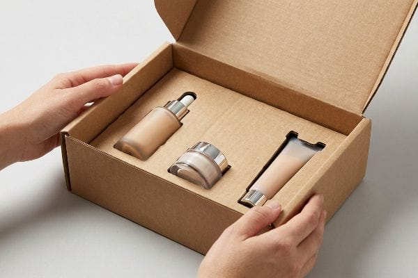 Beauty Box Packaging Insert Brown corrugated beauty subscription box, opened by hands, reveals a custom die-cut insert protecting a foundation dropper, jar, and tube.