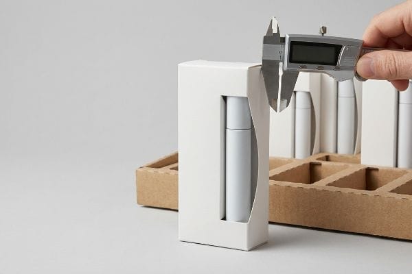 Measuring Cosmetic Box Fit Digital caliper measures a white cosmetic retail box, ensuring precise fit and preventing cylinder bulge in a cardboard display.