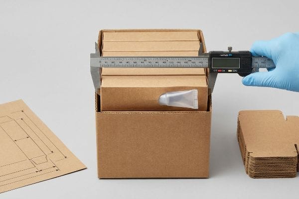 Brown kraft paper boxes are measured by a blue-gloved hand using a Ramtech digital caliper, showing a cosmetic tube and packaging template.