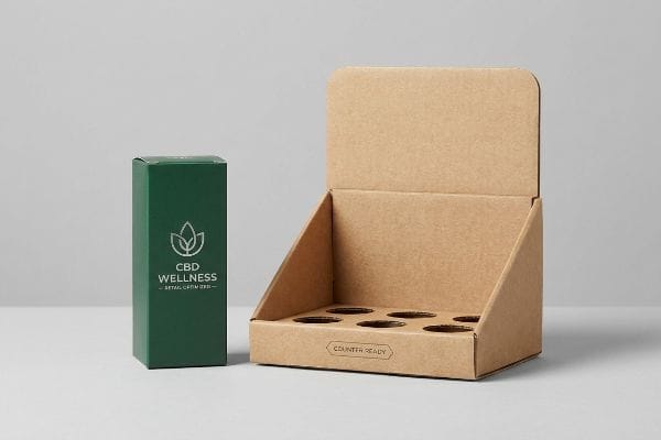 CBD-Wellness-Thekendisplay Eine dunkelgrüne CBD-Wellness-Box steht neben einem leeren, braunen Thekenaufsteller aus Pappe mit sechs Produktfächern.