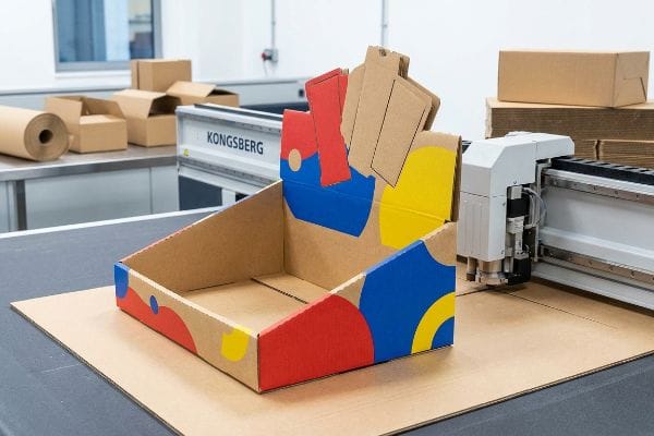 Custom cardboard retail display with red, blue, yellow abstract shapes, on a Kongsberg CNC cutting table.