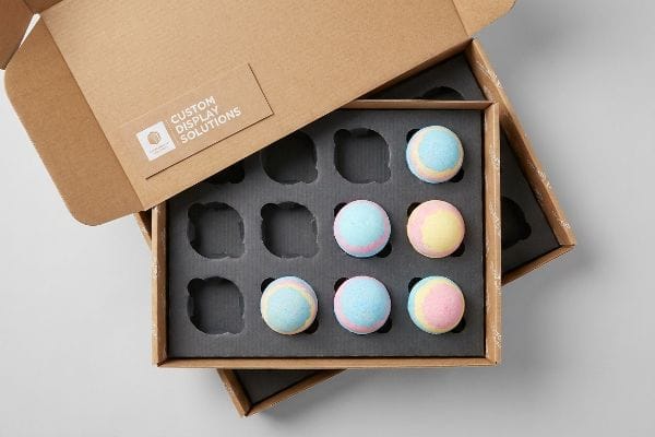 Bath Bomb Display Packaging An open brown corrugated box reveals a dark gray E-flute insert holding six colorful bath bombs, with a "Custom Display Solutions" label on the lid.