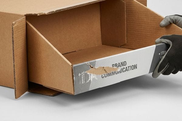 Corrugated retail tray with 'Brand Communication' text and a tear, being pulled from a master carton by a gloved hand.