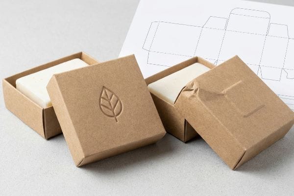 Two open kraft paper soap boxes, one with an inward debossed leaf logo, another slightly crumpled, with a packaging dieline template.
