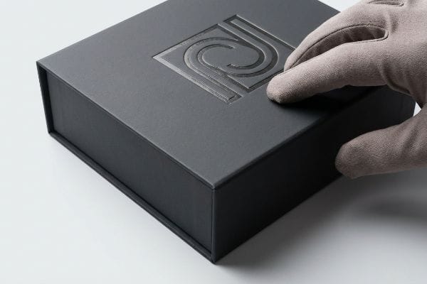 Tactile Debossed Gift Box Dark gray gift box with a debossed, stylized 'P' logo, touched by a gloved hand, showcasing premium tactile packaging.