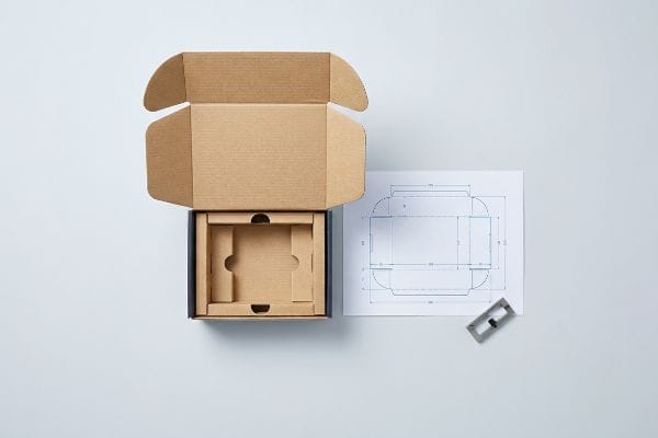 An overhead view shows an open brown corrugated die-cut box with an internal insert, alongside a technical blueprint and a metal die-cutting tool.