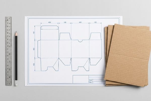 Cosmetic Box Dieline Design The process of cosmetic box structural engineering is shown with a dieline blueprint, corrugated cardboard, ruler, and pencil on a gray background.