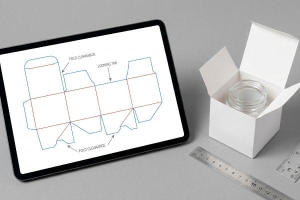 A tablet displays a structural cosmetic packaging dieline with 'Fold Clearance' and 'Locking Tab' details, next to an open white box holding a clear glass jar and a metal ruler.