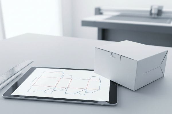 Dieline Packaging Production A modern workspace shows a tablet with a dieline template, a white cardboard box, and a cutting machine in the background, illustrating packaging production.