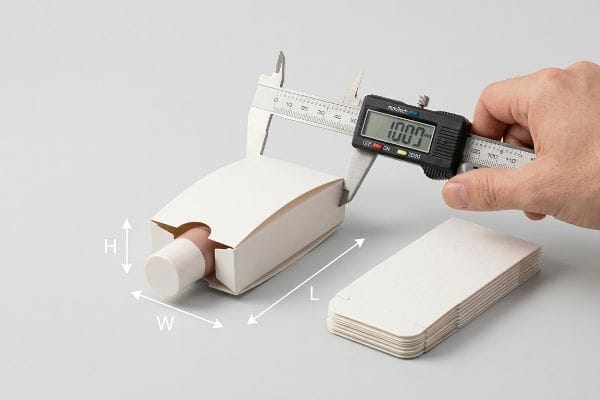 Hand measuring 100.0mm length of a white paperboard box with a cosmetic bottle using a digital caliper, showing box dimensions.
