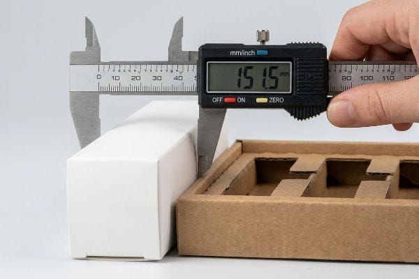 Caliper Measuring Carton Width Hand measuring a white carton's width with a digital caliper showing 151.5 mm, beside a brown corrugated tray.