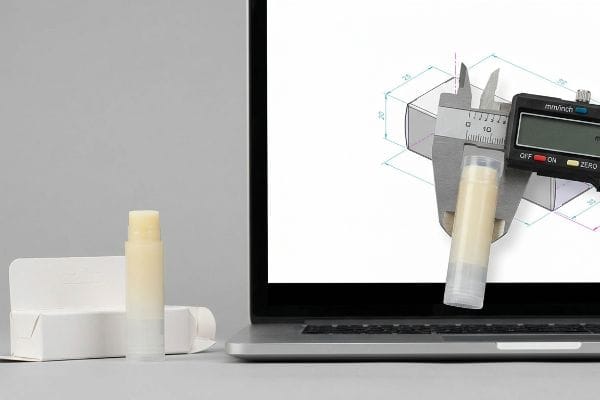 デジタルノギスで、CAD図面が表示されたノートパソコンの画面上でリップクリームのチューブを測定している。近くには、実際のリップクリームと白い紙製のカートンが置かれている。.
