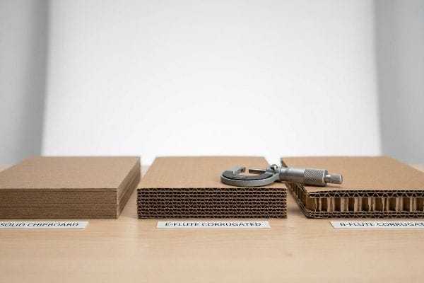 A micrometer rests on E-Flute Corrugated cardboard, comparing it with Solid Chipboard and B-Flute Corrugated materials on a wooden table.
