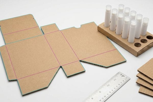 マゼンタ色の折り目が入ったEフルート段ボールの包装を広げたところ、白いリップクリームのチューブが入った型抜きトレイと定規が添えられている。.