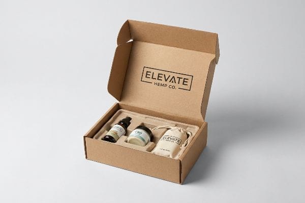 An open corrugated mailer reveals Elevate Hemp Co. CBD products, including a glass tincture, a jar, and a drawstring bag, nestled in a protective insert.