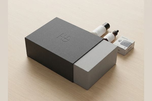 Partially open dark grey sleeve box revealing cosmetic bottles and a small product box, featuring an embossed HG logo.