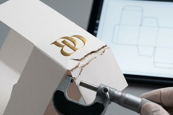 White paperboard packaging with a gold foil embossed BD logo shows a cracked fold, measured by a micrometer, illustrating structural failure.
