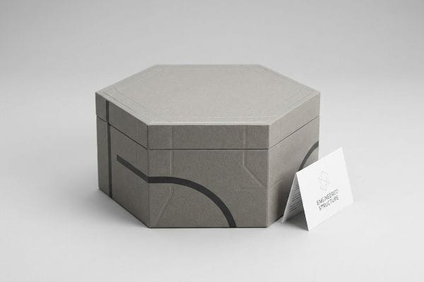 Engineered Hexagonal Box Gray hexagonal rigid box featuring black geometric lines and a subtle embossed lid pattern, positioned next to a white folded card displaying 'Engineered Structure' and a stylized geometric logo.