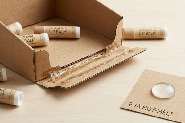 Brown corrugated cardboard display box with organic lip balm tubes, showcasing a clear EVA Hot-Melt adhesive sample and applied glue lines, highlighting solvent-resistant packaging.