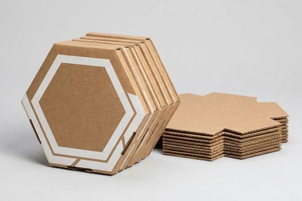 Arranged on a light background, a stack of custom hexagon boxes with white printed outlines stands beside flat-packed corrugated cardboard die-cuts.