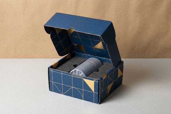 Ein offener, individuell gestalteter Versandkarton in Blau mit goldenen geometrischen Mustern, der über eine schwarze Schaumstoffeinlage zum Schutz eines zylindrischen Produkts verfügt.