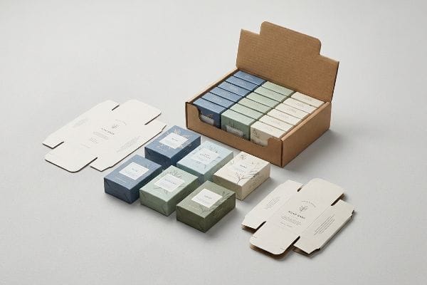 Flat-packed SBS board soap bar cartons and assembled folding cartons in blue, green, and cream, alongside a corrugated PDQ display tray filled with more soap boxes.