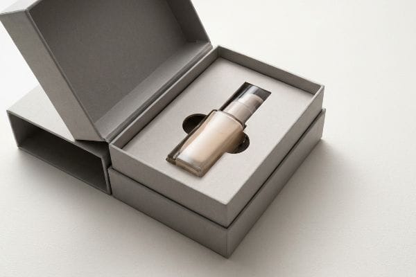 A light beige foundation bottle is securely held within a custom die-cut paper insert inside an open grey rigid cosmetic packaging box, showcasing a suspension void.