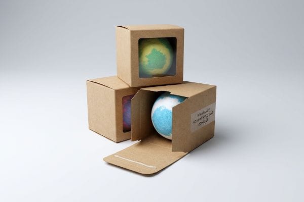 Bath Bomb Packaging Adhesives Kraft paperboard boxes display colorful bath bombs, with one box open revealing a blue bath bomb and a label: "Fragrance Solvent Resistant Adhesive."