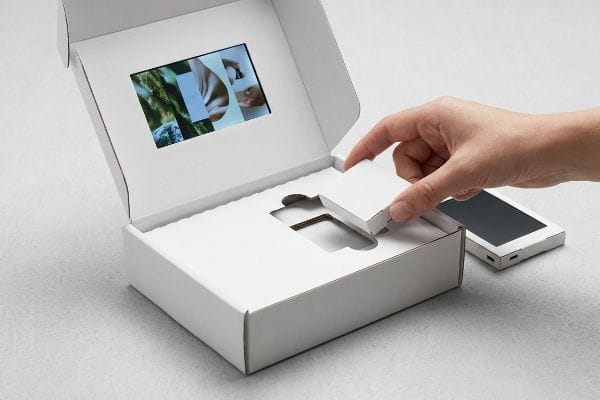 Caixa de papelão ondulado branco com uma tela de vídeo LCD exibindo imagens, enquanto uma mão insere um módulo de encaixe por fricção, separando os componentes eletrônicos do papelão.