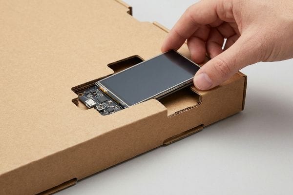 Instalación del bloqueo por fricción de la pantalla Una mano introduce una pantalla electrónica en una cavidad de cartón troquelada con cierre por fricción, dejando al descubierto una placa de circuito para el embalaje interactivo.