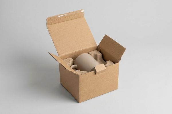An open brown cardboard box reveals a beige mug nestled in protective, recyclable molded pulp inserts, showcasing frustration-free packaging.