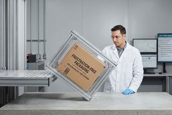 Frustration-Free Packaging Test A brown cardboard box, labeled "Frustration-Free Packaging" and "ISTA 6-Amazon.com-SIOC Certified", is being drop-tested by a technician in a lab.
